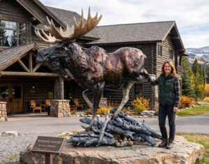 Life-size bronze bull moose monument by Eric Thorsen, running over bronze logs on a stone boulder base in front of a log lodge