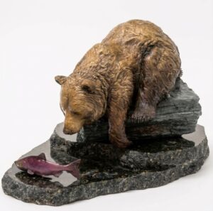 Bronze sculpture of a grizzly bear crouched on a dark stone ledge, looking down at a salmon on the base below