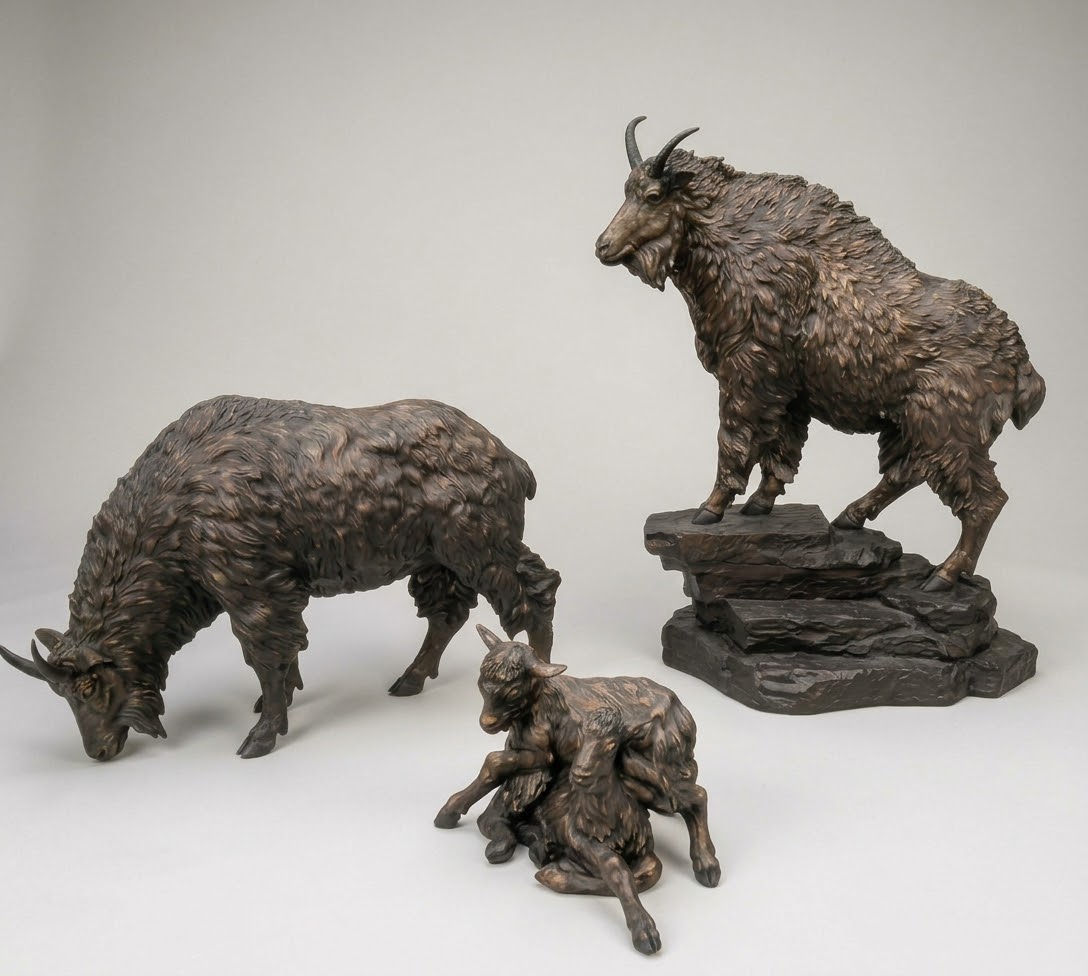 Bronze sculpture group of a mountain goat family: an adult goat standing on a rocky base, a second adult grazing and a kid lying down, on a gray background